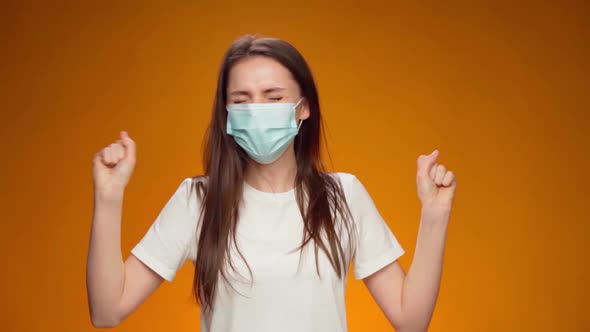 Woman in Medical Mask on Face is Very Happy and Claps Her Hands Against Yellow Background alt