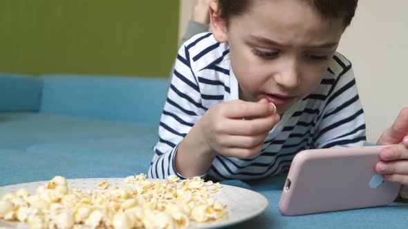 The Child Eats Popcorn Lying on the Sofa in the Room and Spending Time on the Internet Using a alt