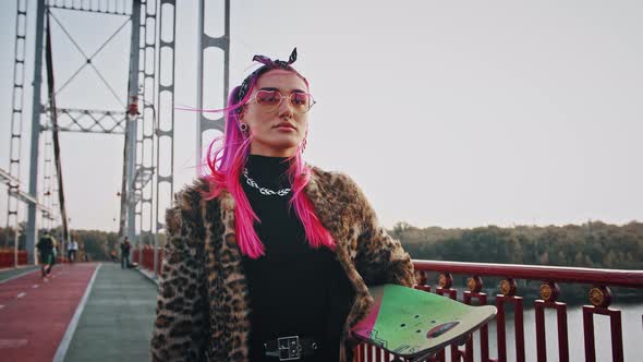 Hipster Woman with Pink Hair in Informal Clothes is Holding Skateboard Walking Across City Bridge alt