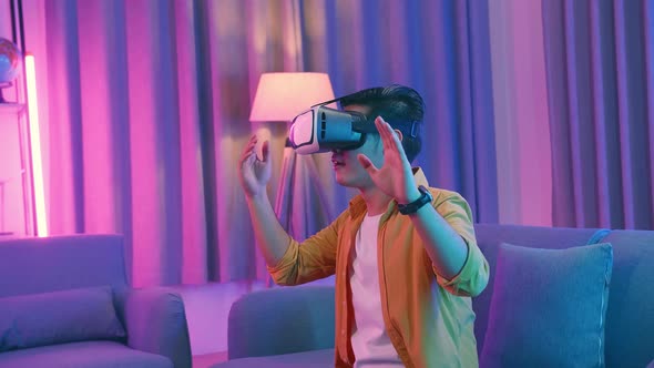 Asian Man Wearing Vr Headset At Living Room, Using Hand Touching And Slide, Cyan And Magenta Colors alt