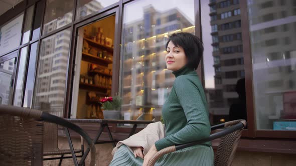 Brunette Woman in Green Sits in a Cafe on the Street Looks at the Camera and Smiles