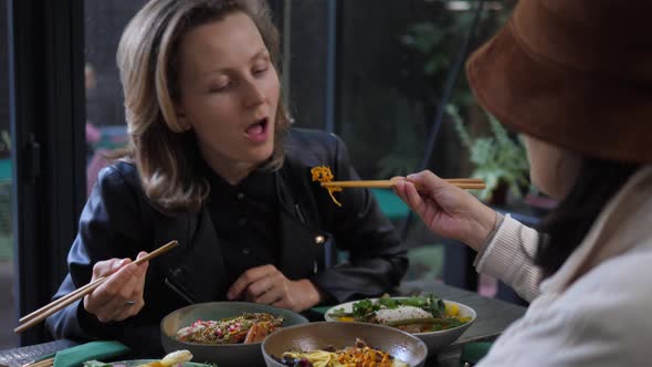 A Girl in a Hat Feeds Blonde Girl with Chopsticks. Sharing Your Food with the Loved Ones alt