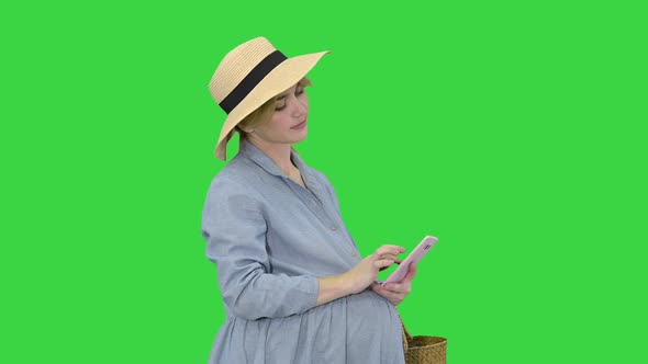 Happy Pregnant Woman in Blue Dress and Hat Using Her Smartphone on a Green Screen, Chroma Key. alt