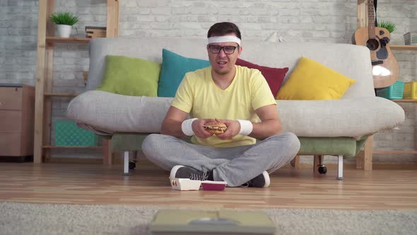 Funny Sporty Man in Sportswear Sitting on the Floor Looking at the Bathroom Scales Eating a Burger alt