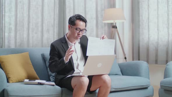 Asian Businessman In Jacket And Shorts Presenting Work Paper On A Laptop At Home alt