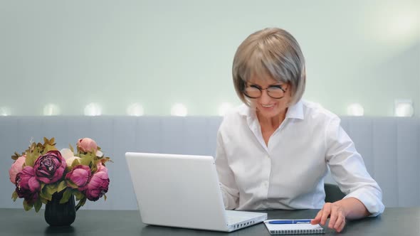 A Happy Beautiful Elderly Woman Enjoys the Good News While Looking at Her Laptop While Sitting at