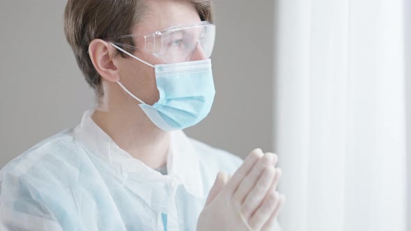 Portrait of Frustrated Young Male Doctor in Covid Face Mask Praying Standing at Window Indoors alt