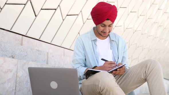Indian Student Man in Traditional Turban Sitting Outdoors and Using Laptop for Learning on Distance