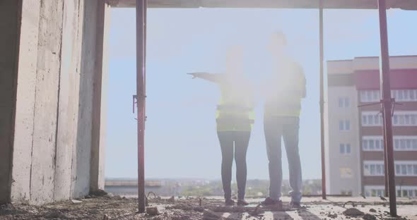Back View Young Foreman and Engineer Checking the Blueprint and Talking About Construction Project alt