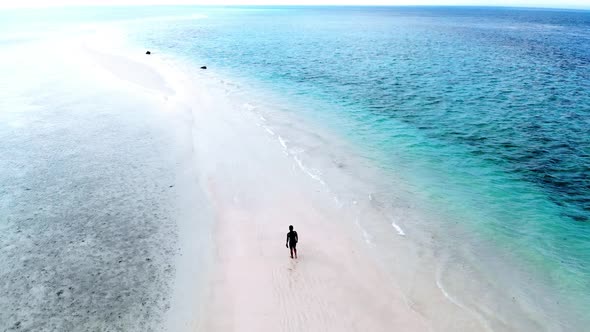 Cinematic aerial clip of a young man walking at the beach with blue water