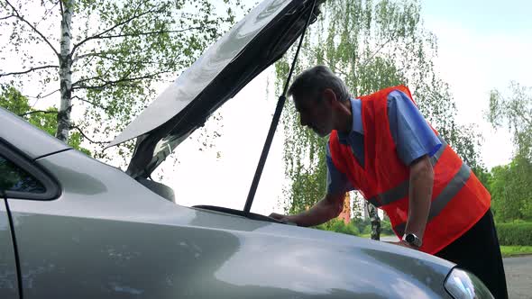Senior Man Wears Warning Vest and Controls Engine of the Car - Trees in the Background alt