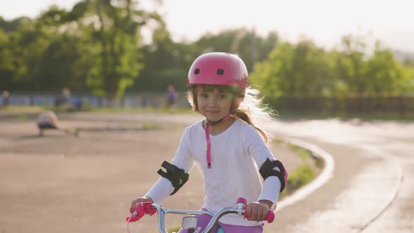 Cute Light Hair Little Girl in Pink Helmet in Elbow and Knee Pads Rides a Bicycle at the Stadium alt