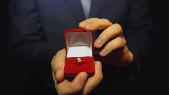 A young man in the blue suit giving a ring with a diamond in the red box