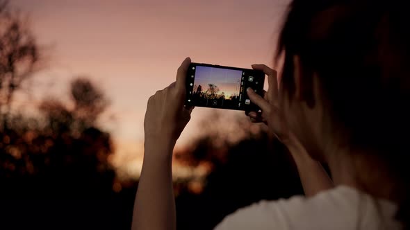Young happy asian girl hold mobile phone relax cheerful take photo peaceful picture of sunset.