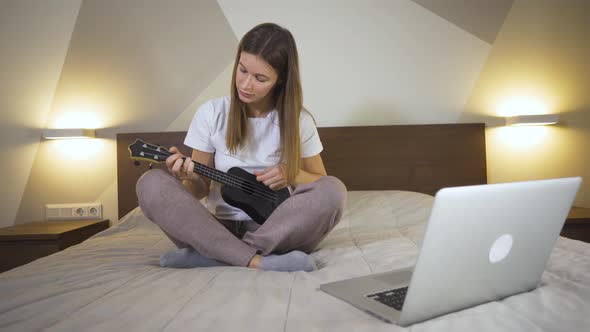Young Woman Playing The Ukulele In House On Bed And Using Laptop in Quarantine. Distance Learning