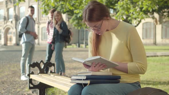 Concentrated Redhead Girl in Eyeglasses Reading Book As Blurred Groupmates Laughing at Her at the alt