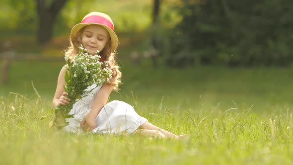 Baby Sits on a Glade in a Sarafan and Hat, Holds Wild Flowers and Smiles alt