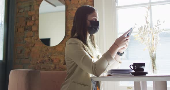 Caucasian female customer wearing face mask, sitting at table, using smartphone alt