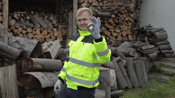 Lumberjack in Reflective Jacket. Man Woodcutter Show Ok Sign. Sawn Logs, Firewood Background alt