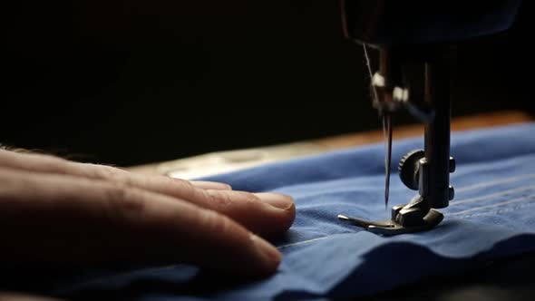 Seamstress Sews With A White Thread On A Sewing Machine A Blue Fabric On A Dark Background