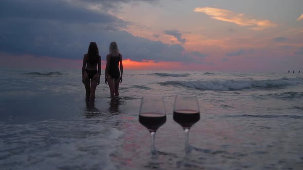 Two women walking in sea in background of two glasses of wine at sunset. alt