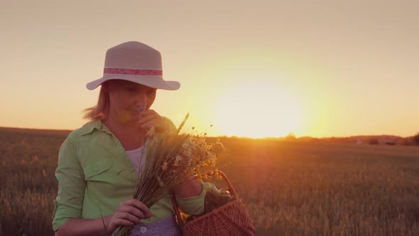 Woman in a Hat with a Bouquet of Wild Flowers Walking Around the Field at Sunset alt