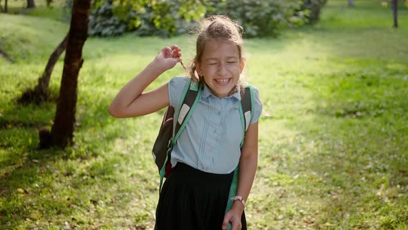 A Pretty Curly Blonde Schoolgirl Looks at the Camera and Smiles. The Child Rejoices After School and alt
