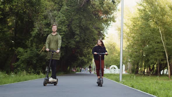 Man and Woman Ride Electric Scooters Along Alley in Public Park alt