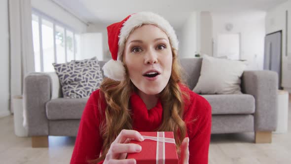 Surprised woman in Santa hat opening a gift box while having a video call on laptop at home alt