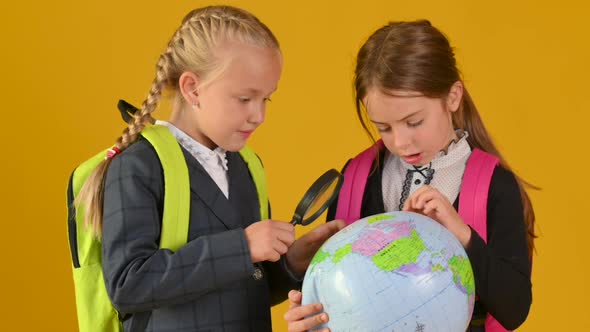 Two cute little schoolgirls in a school uniform with backpacks are holding and learning a globe