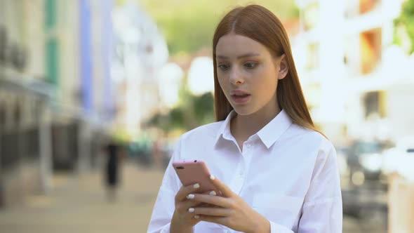 Young Redhead Female Scrolling Social Network on Smartphone, Shocked by News alt