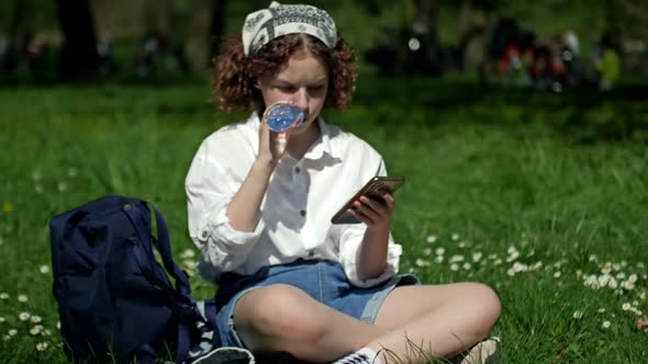 Cute Teenage Girl Sits on the Green Grass in the Park Drinks Water From a Plastic Bottle and Looks