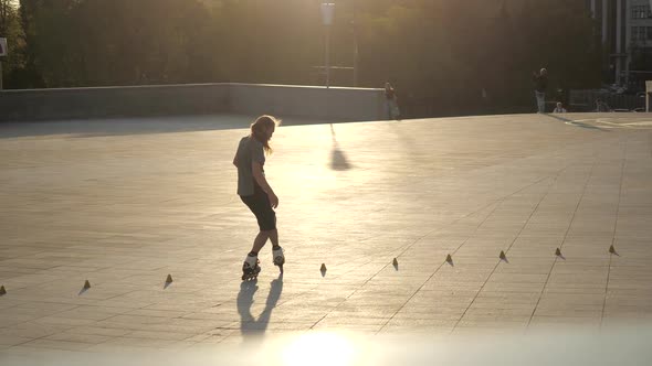 Young Long-haired Man Roller Skater Is Dancing Between Cones in the Evening in a City Square at alt