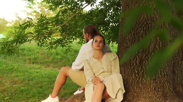 Young Couple Love Sitting with Their Backs to Each Other Under Tree in the Park