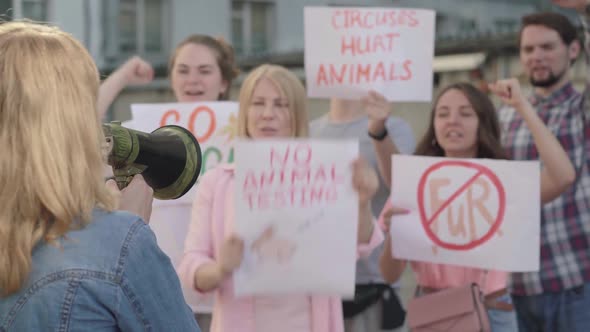 Back View of Young Caucasian Woman Shouting Through Megaphone with Blurred People Supporting alt