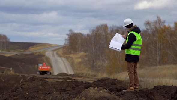 The Worker Looks at the Drawings on the Background of the Road Under Construction alt