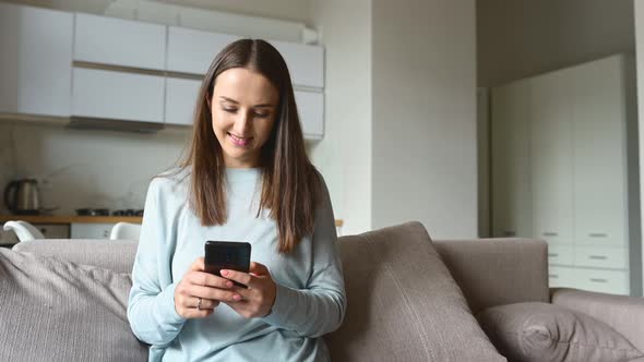 Serene Young Woman Holds Smartphone Sitting on the Couch at Home alt
