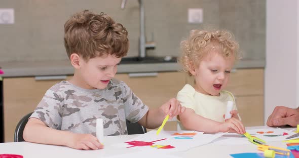 Small Girl and a Small Boy are Making Applications with the Coloured Paper and Glue and Their Father alt