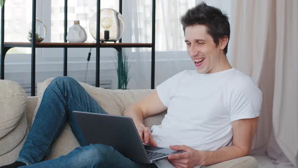 Caucasian Millennial Cheerful Business Man Adult Lies Sitting on Couch at Home Looking at Laptop alt