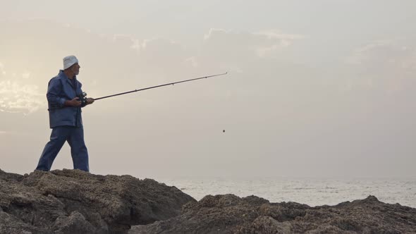 Old fisherman standing on sea side rocks and fishing against the sunset alt