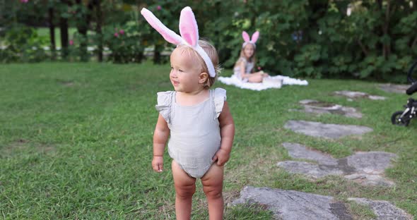 Two Children Wearing Bunny Ears When Pick Up Painted Easter Egg Hunt In Garden or Park alt