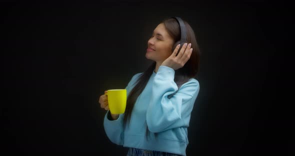 Woman in a Blue Sweater Enjoys Music with Headphones Drinks From a Yellow Mug