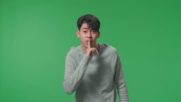A Smiling Asian Man Saying Shh While Standing In Front Of Green Screen Background alt