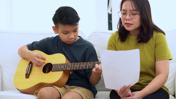 Asian mother embraces son, Asian boy playing guitar and mother embrace on the sofa alt