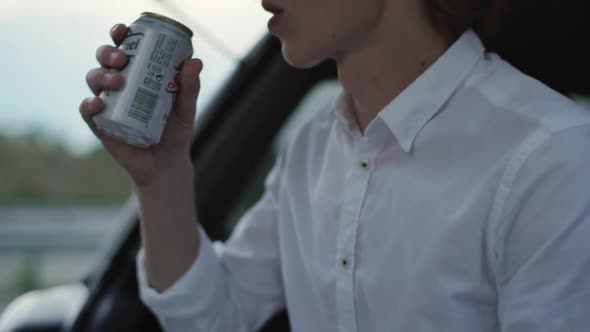 Caucasian Guy in Good Mood Making Pause Sitting in Car on Road During Sunset Drinking Beer From Can alt