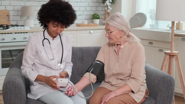 African American Woman Doctor Checking Blood Pressure of Senior Woman While Sitting on Couch at Home alt