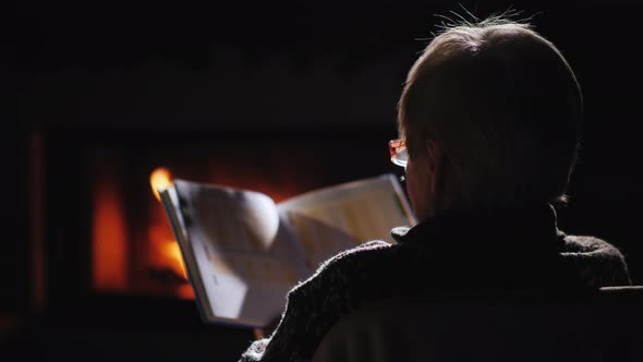 An Elderly Woman in Glasses Reads a Book By the Fireplace alt