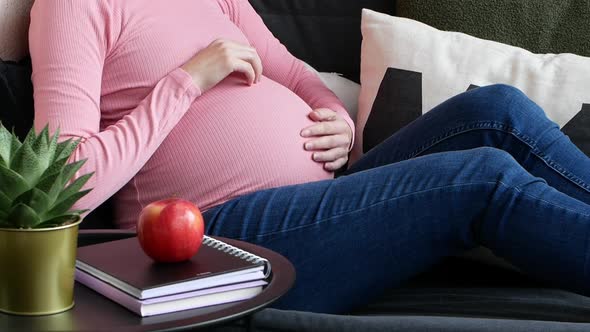 A pregnant woman stroked her belly with her hands while sitting on the couch.