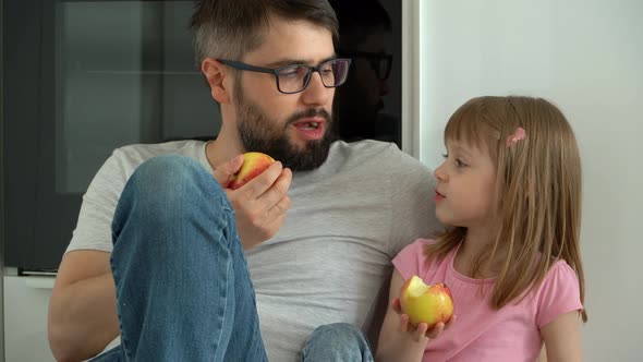 Father and Daughter Sit on Floor at Home in Kitchen and Eat Ripe Apples