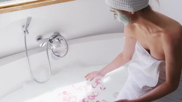 Woman wearing face pack spreading rose petals in the bathtub alt